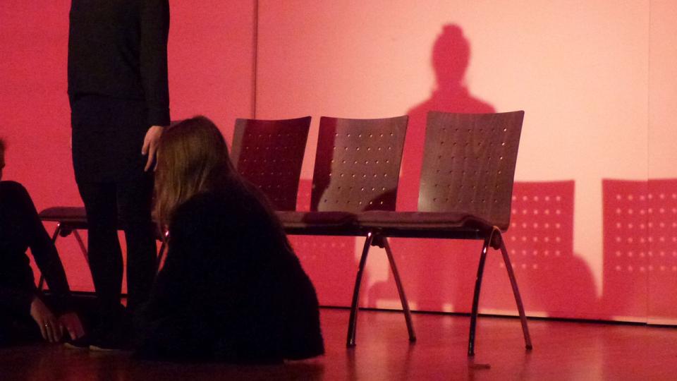 Eine Schauspielerin sitzt vor drei Stühlen mit dem Rücken zur Kamera