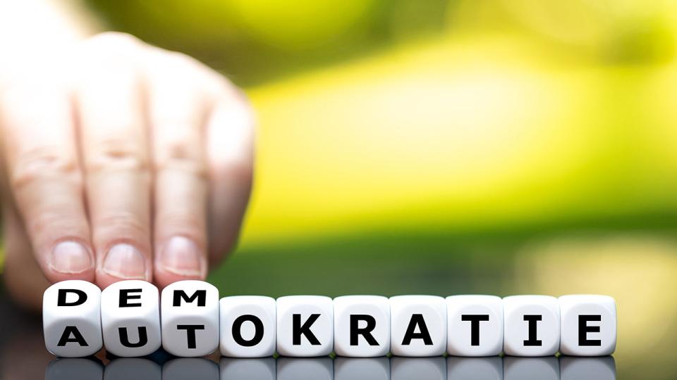 Die Hand würfelt und verwandelt das deutsche Wort „Autokratie“ in „Demokratie“.