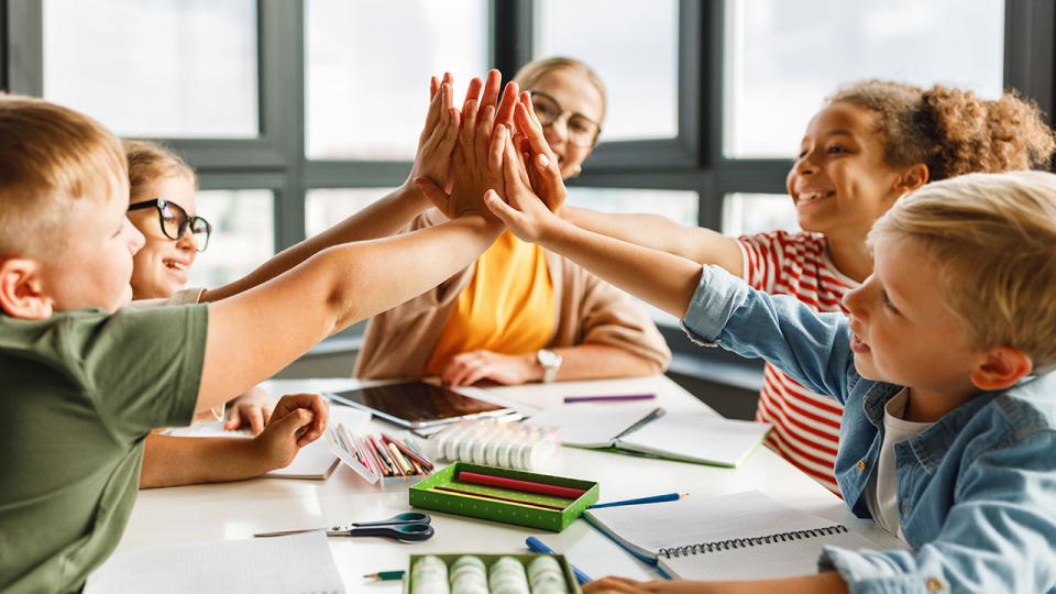 Lehrer und fröhliche Schüler feiern den erfolgreichen Abschluss einer gemeinsamen Schularbeit in einem hellen Klassenzimmer