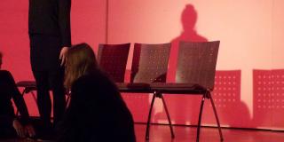 Eine Schauspielerin sitzt vor drei Stühlen mit dem Rücken zur Kamera