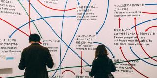 Zwei Menschen stehen vor einer Wand, die mit asiatischen Schriftzügen und Verbindungspfeilen vor diesen erscheint