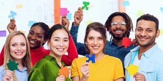 Eine Gruppe von Menschen unterschiedlicher ethnischer Herkunft. Jeder hält ein andersfarbiges Puzzleteil in der Hand.