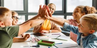 Lehrer und fröhliche Schüler feiern den erfolgreichen Abschluss einer gemeinsamen Schularbeit in einem hellen Klassenzimmer
